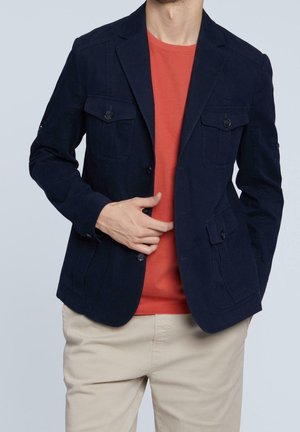 Hombre con una americana azul marino con bolsillos con botones sobre una camiseta naranja y pantalones beige, ajustándose la parte delantera de la americana con la mano.