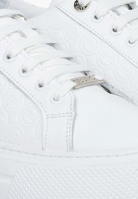 Witte sneaker met een reliëf rond patroon, gladde leermateriaal, witte veters en een gestructureerde rubberen zool. Metalen logo accent nabij de veters.
