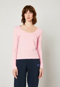 T-shirt a maniche lunghe rosa chiaro con scollo a V in tessuto morbido e elasticizzato. Design semplice con vestibilità aderente, indossato con pantaloni blu scuro con un logo.