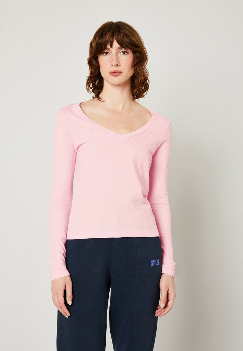 T-shirt a maniche lunghe rosa chiaro con scollo a V in tessuto morbido e elasticizzato. Design semplice con vestibilità aderente, indossato con pantaloni blu scuro con un logo.