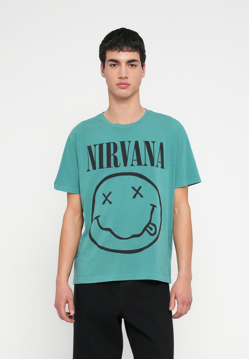 Τυρκουάζ βαμβακερό μπλουζάκι με μεγάλο μαύρο κείμενο "NIRVANA" και ένα γραφικό με χαμογελαστό πρόσωπο με σταυρωμένα μάτια και γλώσσα έξω. Κοντά μανίκια.