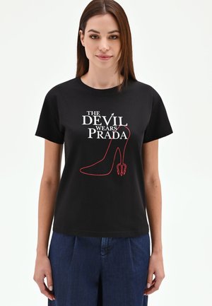 Donna che indossa una t-shirt nera con la scritta "The Devil Wears Prada" e il contorno di una scarpa con tacco rosso dotata di coda di diavolo e forcone.