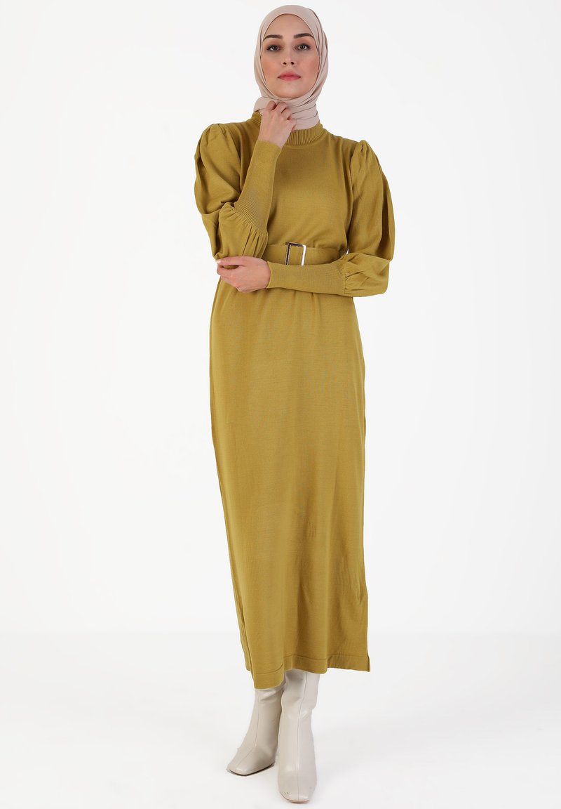 Largo vestido verde mostaza con mangas largas abullonadas, cuerpo ajustado y un cinturón que acentúa la figura. Llevado con botas de tacón a media pantorrilla de color crema.