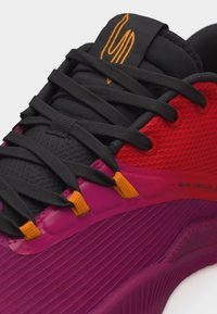 Nærbilde av en lilla og rød sportssko med svarte skolisser tredd gjennom oransje hull, med en svart tunge med oransje logo.