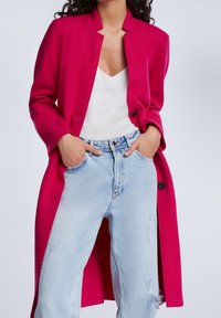 Kirkkaan vaaleanpunainen pitkä villakangastakki, jossa on lovikaulus, kaksi nappia ja sivuhalkiot, yhdistettynä vaaleisiin kulutettuihin sinisiin farkkuihin.