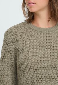 Strickpullover in Olivgrün mit strukturiertem Muster, Rundhalsausschnitt und gerippten Bündchen. Nahaufnahme des Stoffdetails.