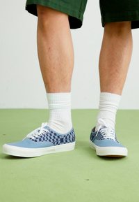 Veterschoenen met een blauwe stoffen bovenkant en een dambordpatroon. Witte rubberen zool, gestyled met witte crew-sokken en groene shorts.