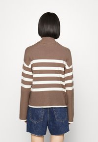 Person med kort svart hår som bär en brun och vit randig tröja samt blå denimshorts, stående med ryggen mot en enhetlig bakgrund.