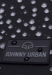 Fekete textúrált anyag vízcseppekkel; egy szürke gumibetét található rajta, amelyen dombornyomott "JOHNNY URBAN" felirat és logóelemek láthatók.