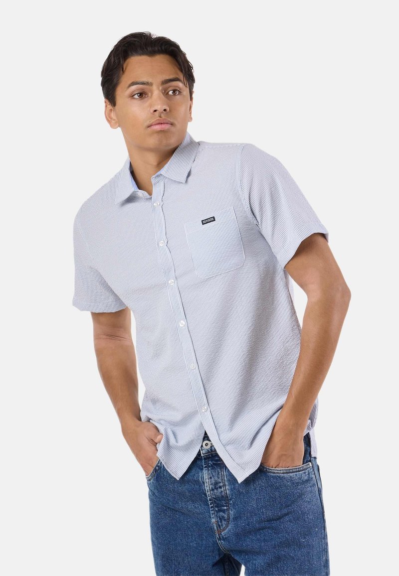 Jeune homme portant une chemise à manches courtes bleu clair à rayures avec une poche avant et un jean bleu, debout les mains dans les poches.