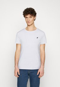 White cotton crew-neck t-shirt with short sleeves, featuring a small black bird logo on the left chest. Worn with blue denim jeans.