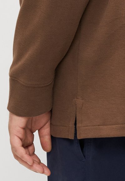 Bruine lange mouwen shirt met een geribbelde textuur, zijventilatie en een relaxte pasvorm. Hand rust nabij de zoom tegen de marineblauwe broek.