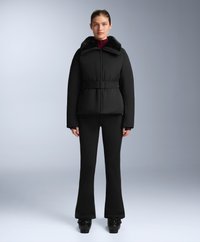 Žena odjevena u crnu podstavljenu jaknu s krznom na ovratniku i remenom, crne trapezaste hlače i crne cipele, stoji na sivoj pozadini.