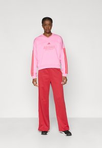 Rózsaszín pulóver piros kiegészítőkkel és logóval, piros nadrággal párosítva, amelyen fehér oldalsávok vannak. Simább anyag, laza szabás.