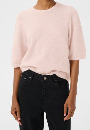 Pull rose clair à manches courtes en matériau doux et moelleux avec un col rond, associé à un jean noir taille haute avec des attaches métalliques.