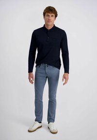 Man staat, gekleed in een donkerblauwe poloshirt met lange mouwen, lichtblauwe broek en witte sneakers, tegen een effen lichtgrijze achtergrond.