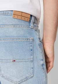 Jeans de mezclilla azul claro con una etiqueta rectangular de cuero marrón claro en la cinturilla trasera, etiquetada como "J.E.A.N.S.", y un pequeño logo de marca en el bolsillo.