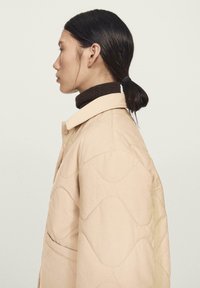 Beżowa pikowana kurtka o miękkiej fakturze, z kołnierzem i zapięciem na guziki, noszona na brązowym golfie.