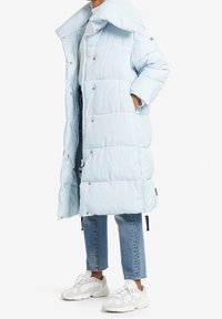 Ljusblå puffjacka med hög krage, tryckknappar och sidfickor, kombinerad med korta denimjeans och vita sneakers.