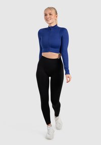 Blauwe gecropte zip-up top met lange mouwen, ribbelstructuur en hoge kraag, gecombineerd met zwarte leggings en witte sportschoenen.