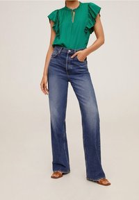 Grøn bluse med rynker og nøglehulsudskæring, parret med højtaljede, vidde jeans i blå og brune sandaler. Neutral baggrund.