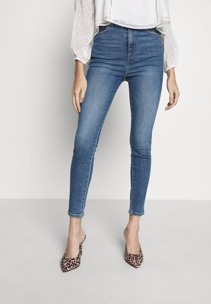 Femme portant un jean skinny bleu taille haute, une blouse blanche à manches longues avec manches transparentes à pois, et des talons pointus imprimé léopard.