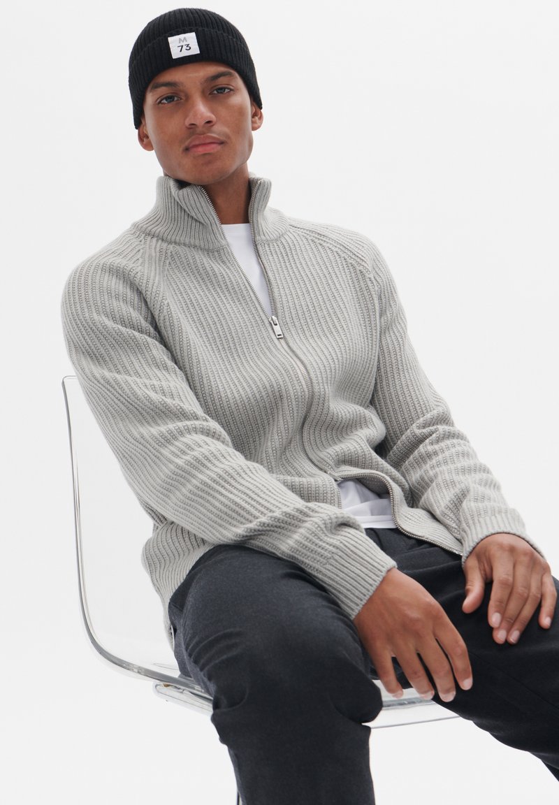 Helles graues, geripptes Zip-Up-Sweatshirt mit hohem Kragen, lange Ärmel und lockerer Passform, getragen über einem weißen T-Shirt mit dunklen Hosen.