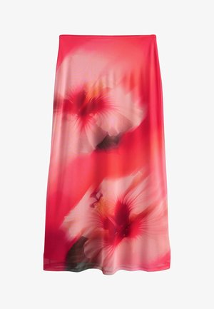 Mujer con un vestido largo y ceñido de color coral con grandes estampados florales, de pie con las manos apoyadas en las caderas y calzando sandalias marrones.
