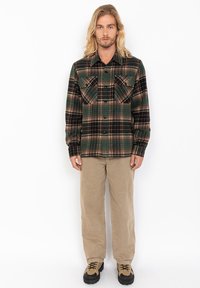 Sort og grønternet flannel skjorte med to brystlommer, knappelukning og lange ærmer, parret med beigefarvede løstsiddende bukser og brune støvler.