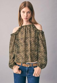 Ung kvinde med lige, lysebrunt hår iført en kold-skulder leopardmønsteret bluse, blå jeans og en tanfarvet bælte foran en ensfarvet baggrund.