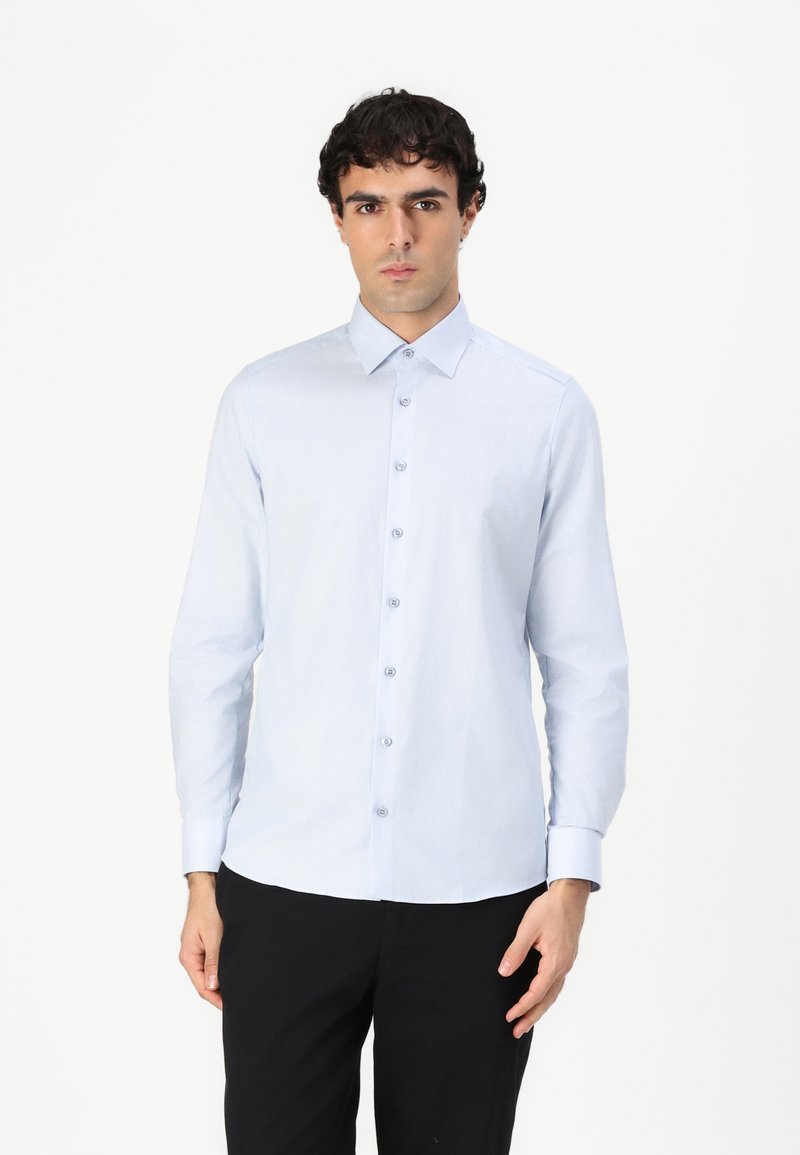 Chemise à manches longues bleu clair avec col à boutons, dotée de boutons sur le devant et d'une texture tissée lisse, associée à un pantalon sombre.