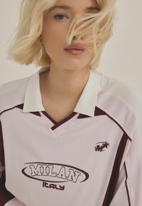 Růžový a bílý sportovní dres s kaštanovými akcenty. Vybaven límečkem a grafikou "MILAN ITALY" na přední straně. Lehce materiál.