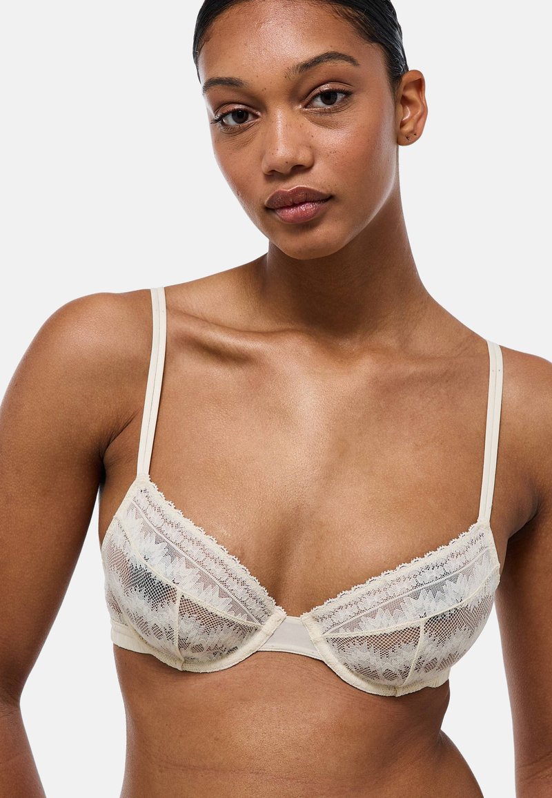 Soutien-gorge en dentelle beige avec des panneaux transparents, des bonnets triangulaires, des bretelles ajustables et des détails festonnés le long des bords.