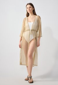 Cubierta dorada y transparente con mangas tipo kimono, abertura frontal y cintura ajustada. El tejido tiene una textura brillante y cae hasta la mitad de la pantorrilla.