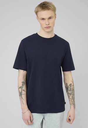 Jeune homme aux cheveux blonds courts portant un t-shirt bleu marine uni, un pantalon clair rayé et des tatouages visibles sur les deux avant-bras.