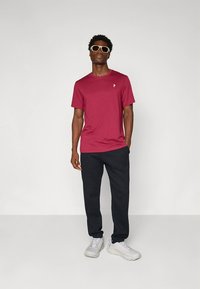 Dunkelrotes T-Shirt mit kurzen Ärmeln und kleinem Logo auf der Brust, kombiniert mit schwarzen Jogginghosen und hellgrauen Sneakern. Der Hintergrund ist einfarbig.