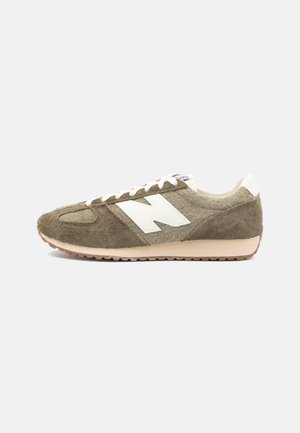Niski brązowy zamszowy sneaker z białymi sznurowadłami i dużym białym logo "N" po boku, beżowa podeszwa i biała zawieszka na pięcie.