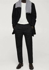 Personne portant un manteau noir, une chemise blanche, un pantalon noir, des mocassins foncés et une écharpe en laine grise drapée sur une épaule. Mains dans les poches.