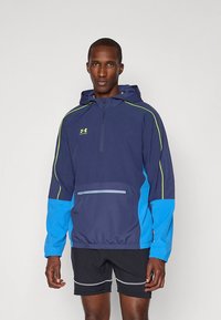 Tumesinine tuulejakk kapuutsiga, kontrastse siniste varrukatega, neoonroheliste detailidega ja ees tõmblukuga taskuga. Kerge ja sportliku lõikega.