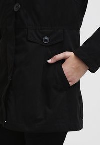 Main placée dans la poche latérale d'une veste noire avec une poche à rabat boutonnée au-dessus sur un fond clair.