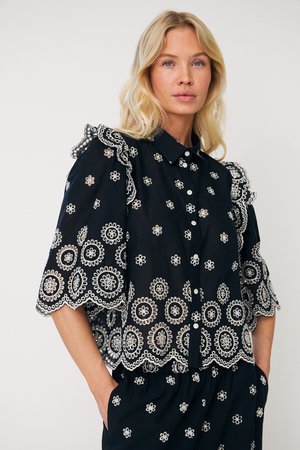 Blonde Frau in schwarzer Bluse mit weißer Blumenstickerei und Rüschen an den Schultern, stehend mit den Händen in den Taschen der passenden bestickten Hose.