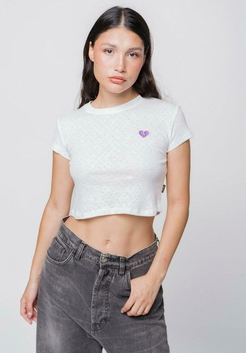 Camiseta blanca corta con una textura sutil y un parche de corazón morado en el lado izquierdo del pecho, combinada con jeans grises de cintura alta.