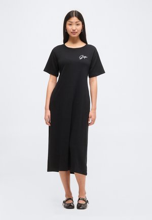 Kobieta stoi, nosząc czarną midi sukienkę w stylu T-shirt z logo "Gap" na piersi oraz czarne buty Mary Jane na jednolitym tle.