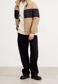 Beżowa i czarna kurtka z zamkiem błyskawicznym, z designem w formie bloków kolorystycznych i elastycznymi mankietami, noszona na kremowej koszuli z długim rękawem i czarnych spodniach.