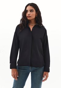 Frau mit langen, welligen Haaren, die ein schwarzes Knopfhemd, eine blaue Jeans, eine goldene Kettenhalskette und goldene Creolen trägt und zur Seite schaut.
