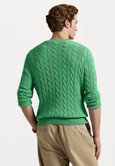 Grønn kabelstrikket genser med ribbestrikket kant og mansjetter, med et teksturert mønster. Båret over beige bukser, som viser bakvinkelen.