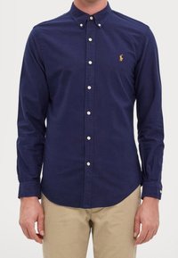 Navy blue button-up shirt in cotton with long sleeves, collar, and a small embroidered logo on the left chest. White buttons are visible.