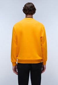 Geel sweatshirt met een ronde halslijn, ribgebreide manchetten en zoom. De achterkant heeft een gladde stof en een eenvoudig ontwerp. Gepaard met zwarte broeken.