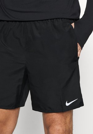 Personne portant un short Nike noir avec une taille élastique, la main dans la poche droite, et un haut noir à manches longues visible au-dessus du short.