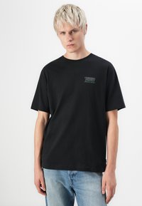Sort bomulds T-shirt med korte ærmer, rund hals og "TURBO Classics" logo i hvid og grøn på venstre brystområde. Båret med lyseblå jeans.